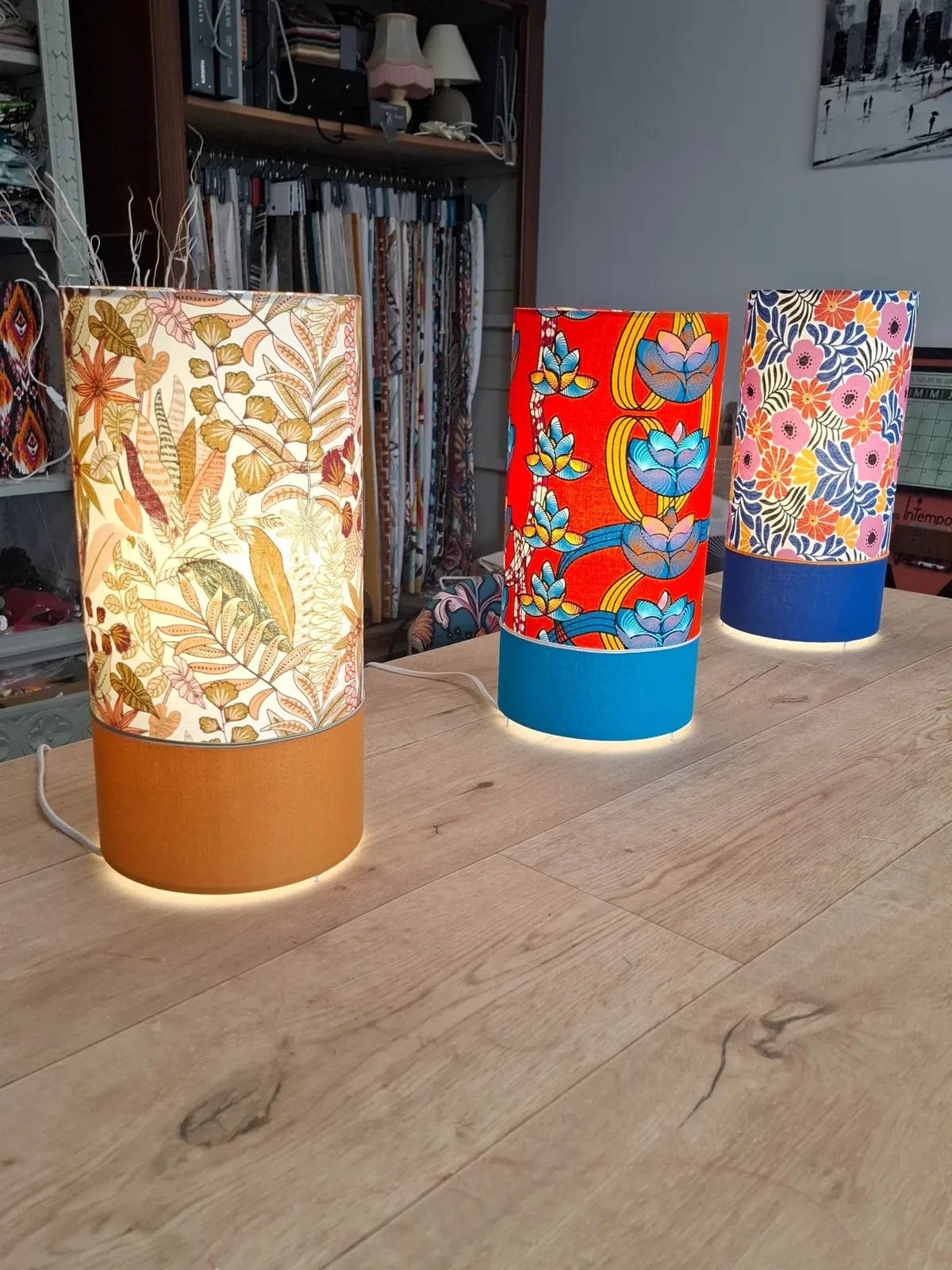 Trois lampes de stage aux tissus feuillage, fleuri rouge et fleuri bleu, allumées sur une table en bois
